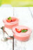 Two glasses of pink strawberry mousse topped with strawberry slices and mint leaves, set on a white wooden table with spoons.
